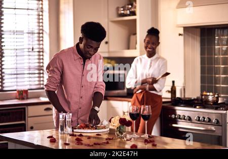 Heute seinen Tag, um sie zu beeindrucken. Eine kleine Aufnahme eines hübschen, romantischen jungen Mannes, der Gemüse hackt und seine Frau im Hintergrund steht. Stockfoto