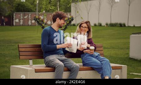 Ein gutaussehender Mann bringt ein Mittagessen für einen Kollegen, der auf einer Bank im Park sitzt. Kaffeepause. Studenten erholsame Zeit. Spaziergang im Freien. Achten Sie auf das Konzept. Stockfoto