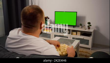 Mann beobachtet Green Mock-up Screen TV, während er abends auf einer Couch zu Hause im Living Room Over the Shoulder Shot sitzt. Stockfoto