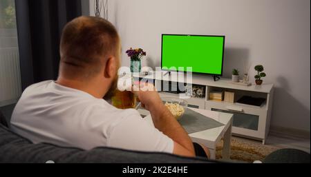 Mann, der zu Hause einen Fernseher mit grünem Bildschirm sieht. Trinken Sie Bier Essen Popcorn. Über der Schulter Rückansicht. Chroma-Taste. Stockfoto