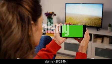 Horizontaler grüner Bildschirm für Copy Space Close Up Chroma Key Mockup. Zurück über die Schulter View Girl sieht sich einen Film auf Couch Home an. Benutzer von Handyhaltetechnologie. Stockfoto