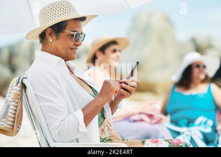 Schicken Sie all unsere Selfies an unsere Kinder. Aufnahme einer reifen Frau, die alleine sitzt und ihr Handy während eines Tages am Strand mit Freunden benutzt. Stockfoto