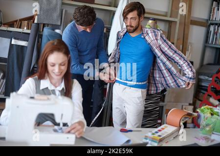 Änderungen vornehmen, während Sie warten. Aufnahme eines jungen Modedesigners, der näht, während ein Kollege im Hintergrund mit einem Kunden zusammenarbeitet. Stockfoto