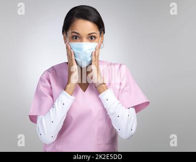 Das kann nicht sein. Ein beschnittenes Porträt einer attraktiven jungen medizinischen Mitarbeiterin, die eine Maske trägt und vor grauem Hintergrund schockiert im Studio aussieht. Stockfoto