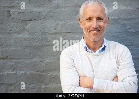 Das ist ein selbstbewusstes Lächeln. Porträt eines reifen Geschäftsmannes, der mit gefalteten Armen an einer Ziegelwand steht. Stockfoto