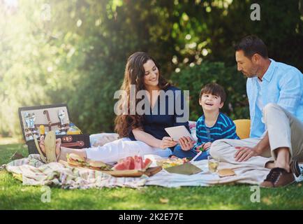 Draußen zu Mittag zu essen ist super. Aufnahme einer jungen Familie mit einem digitalen Tablet während eines Picknicks im Park. Stockfoto