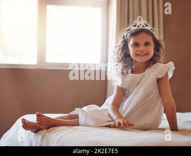 Ist sie nicht nur die süßeste Prinzessin, die du je gesehen hast. Porträt eines entzückenden kleinen Mädchens, das eine Tiara trägt und zu Hause auf einem Bett sitzt. Stockfoto