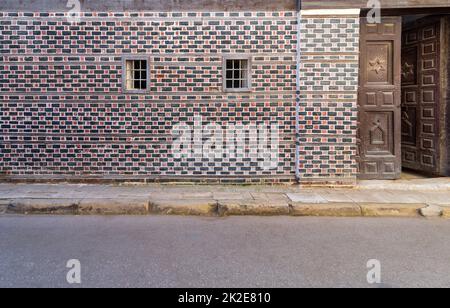 Wand mit schwarzen und roten Ziegeln mit weißer Naht und zwei kleinen Fenstern mit Metallriegeln Stockfoto