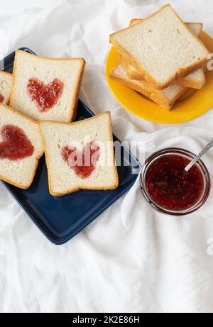 Toast, auf dem das Herz aus Marmelade besteht. Überraschendes Frühstückskonzept im Bett. Romantik für St. Valentinstag. Stockfoto