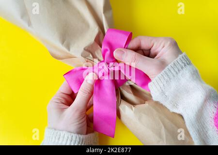 Draufsicht auf weibliche Hände, die ein Blumenstrauß mit einem pinkfarbenen Band und Geschenken auf gelbem Hintergrund binden. Nahaufnahme, Platz für eine Inschrift. Stockfoto