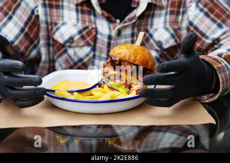 Das Konzept von Fast Food und Essen zum Mitnehmen. Ein Mann in schwarzen Latexhandschuhen ist bereit, einen saftigen Hamburger zu essen, der neben den pommes frites auf einem Metallteller liegt, zusammen mit Käsesauce. Stockfoto