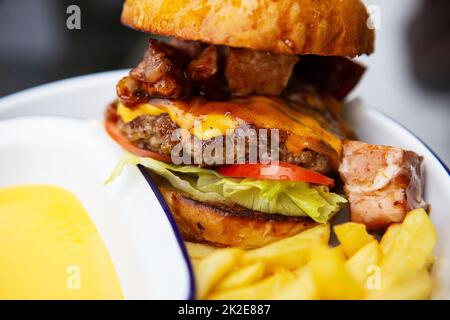Das Konzept von Fast Food und Essen zum Mitnehmen. Ein saftiger Hamburger liegt neben pommes frites auf einem Metallteller zusammen mit Cheddar Cheese Sauce im Hintergrund. Stockfoto