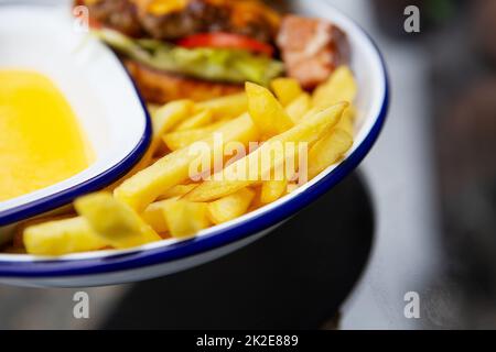 Das Konzept des Fast Food. Pommes frites liegen auf einem Meta-Teller zusammen mit Cheddar-Käsesauce und einem Hamburger im Hintergrund. Stockfoto