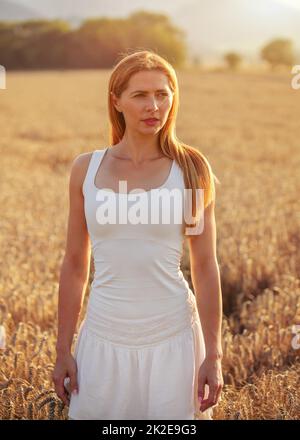 Junge sportliche Frau in weißem Kleid, lange braune Haare, in die Ferne schauen, Sonnenuntergang beleuchtet Weizenfeld hinter ihr. Stockfoto