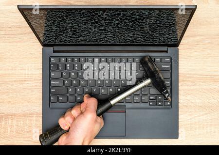 Hand mit einem Hammer und Laptop mit einem kaputten Bildschirm Stockfoto