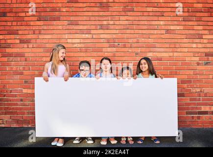 Nun, behalten Sie diesen Raum im Auge. Porträt einer Gruppe von kleinen Kindern, die ein leeres Schild an einer Ziegelwand halten. Stockfoto