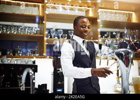 Ich zeige allen meinen Kunden eine gute Zeit. Zugeschnittene Aufnahme eines gut gekleideten Barkeepers, der hinter der Theke steht. Stockfoto