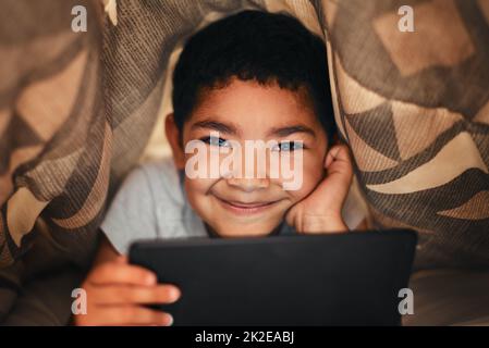 Wie die Schlafenszeit in der Moderne aussieht. Porträt eines entzückenden kleinen Jungen, der ein digitales Tablet benutzt, während er zu Hause unter einer Decke liegt. Stockfoto