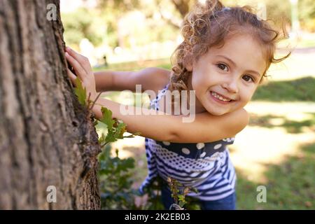 Sie werden mich hier nicht finden. Porträt eines kleinen Mädchens, das sich hinter einem Baum in einem Park versteckt. Stockfoto