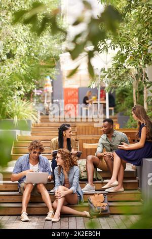 Chillen auf dem Campus zwischen den Klassen. Eine Gruppe junger Studenten, die sich mit technischen Geräten im Freien entspannen. Stockfoto