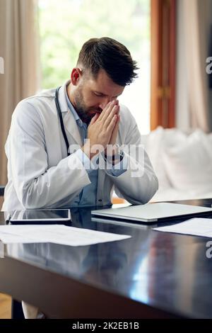 Auf der Suche nach einigen definieren Intervention. Eine kurze Aufnahme eines hübschen männlichen Arztes, der während des Sitzens in seinem Büro betet. Stockfoto