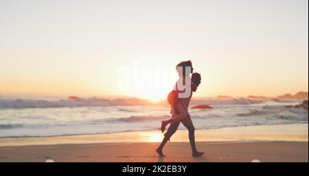 Liefen in den Sonnenuntergang. Ganzkörperaufnahme eines liebevollen jungen Paares, das bei Sonnenuntergang am Strand spazieren geht. Stockfoto