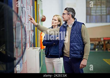 Zwei Manager sprechen zusammen, während sie an einem Pinnwand auf dem Boden der Fabrik stehen. Stockfoto