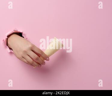 Leere Eisbecher mit Waffelkegel in der Hand einer Frau, die aus einem zerrissenen Loch in einem rosa Papier herausragt Stockfoto