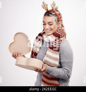 Was ist in der Box. Studioaufnahme einer attraktiven jungen Frau, die eine herzförmige Box öffnet, während sie in weihnachtlicher Kleidung gekleidet ist. Stockfoto