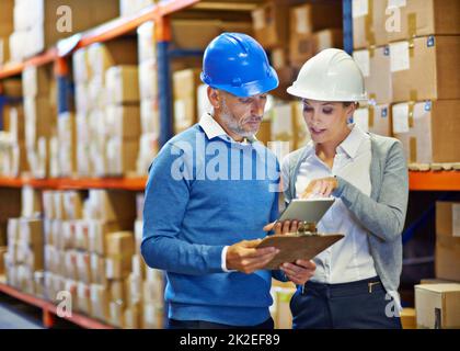 Vergleichen von Notizen zur Genauigkeit. Aufnahme von zwei Personen, die in einem Lagerhaus einen Lagerbestandsscheck machen. Stockfoto