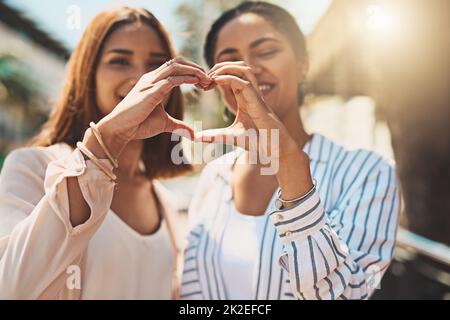 Liebe hält deine Freundschaften stark. Aufnahme von zwei fröhlichen jungen Frauen, die sich tagsüber im Freien eine Herzform bilden und dabei ihre Hände zusammenhalten. Stockfoto