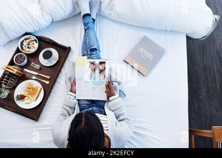 Frühstück im Bett und ein paar gute Bücher. Aufnahmen aus dem Blickwinkel einer unbekannten Frau, die eine Zeitschrift liest und zu Hause auf ihrem Bett frühstückt. Stockfoto