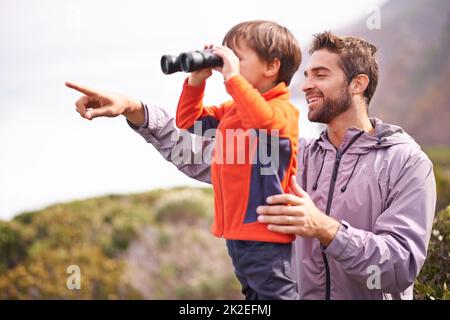 Mit seinem kleinen Jungen die Natur teilen. Aufnahme eines Vaters, der auf etwas zeigt, während sein Sohn durch ein Fernglas auf einer gemeinsamen Wanderung schaut. Stockfoto