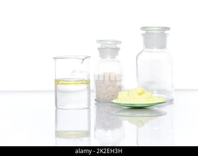 Chemische Inhaltsstoffe auf weißem Labortisch. Schwefelpulver in chemischem Uhrglas neben Öl im Becher und Flakensalz in chemischem Reagenzglas. Seitenansicht Stockfoto