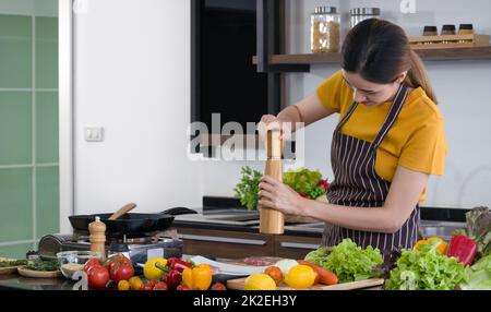 Junge Hausfrau, die Pfeffer auf das Schweinefleisch legt, bevor sie es zu einem Steak kocht. Morgenatmosphäre in einer modernen Küche. Die Küchentheke war voll mit verschiedenen Gemüsesorten. Stockfoto