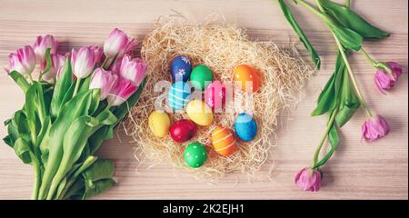 Bunte ostereier im Nest und Tulpen auf dem Holzhintergrund. Stockfoto