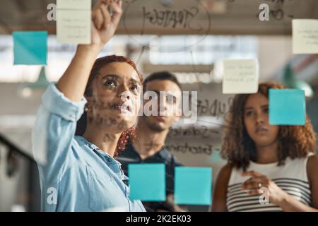 Sie widmen viel Zeit der Perfektionierung ihrer Pläne. Aufnahme einer Gruppe junger Designer beim Brainstorming mit Notizen an einer Glaswand in einem Büro. Stockfoto