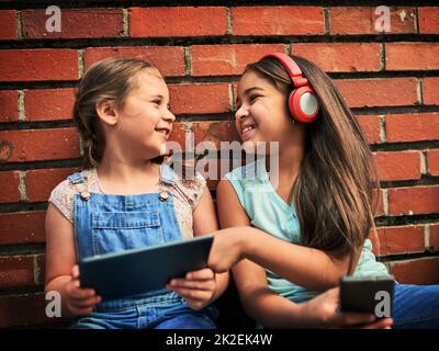 Sie genießen es, lustige Videos zusammen zu sehen. Aufnahme von zwei jungen Mädchen, die digitale Geräte gegen eine Ziegelwand verwendeten. Stockfoto