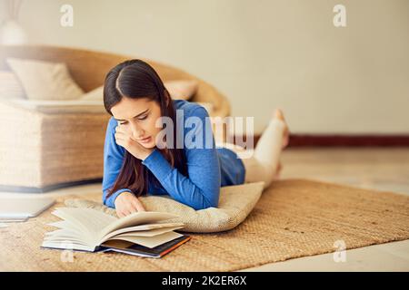 Wirklich in die Erzählung vertieft. Aufnahme einer jungen Frau, die zu Hause ein Buch auf ihrer Lounge-Etage liest. Stockfoto