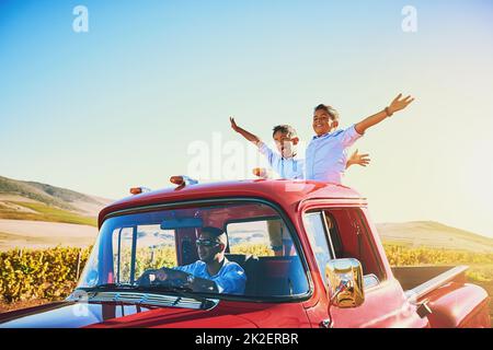 Freiheit gibt es in vielen Formen. Aufnahme einer fröhlichen jungen Familie, die in einem roten Pickup-Truck auf einer Landstraße vor der Tür fährt. Stockfoto