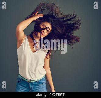 Ihr Haar hat einen eigenen Kopf. Studioaufnahme einer sorglosen jungen Frau, die ihr Haar hält, während es vom Wind geblasen wird, während sie vor einem dunklen Hintergrund steht. Stockfoto