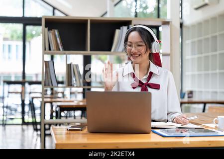 Lächelnde asiatische Frau im Headset winkende Hand, mit Laptop, Blick auf den Bildschirm, Call-Center-Betreiber Beratung Kunden, Schüler lernen Stockfoto