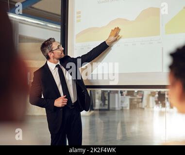 Er kennt seine Zahlen. Aufnahme einer Führungskraft, die eine Präsentation auf einer Projektionsleinwand vor einer Gruppe von Kollegen in einem Sitzungssaal vorführt. Stockfoto