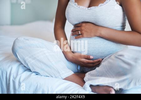 Du verstehst das Leben, wenn es anfängt, in dir zu wachsen. Gekürzte Aufnahme einer unbekannten schwangeren Frau, die auf einem Bett sitzt und ihren Bauch zu Hause hält. Stockfoto