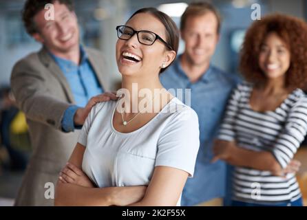 Das Leben ist großartig mit der Unterstützung meiner Kollegen. Junge Geschäftsfrau lacht fröhlich mit unterstützenden Kollegen hinter ihr. Stockfoto