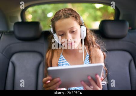 Sehen Sie sich ihre Filme auf dem Rücksitz an. Aufnahme eines jungen Mädchens, das in einem Autositz mit Kopfhörern sitzt und ein digitales Tablet verwendet. Stockfoto