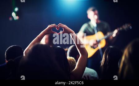 Aus Liebe zur Musik. Eine kurze Aufnahme von Händen einer Frau, die sich nicht wiedererkennen lassen und eine Herzform bilden, während ein Musiker bei einem Konzert in der Nacht auftritt. Stockfoto