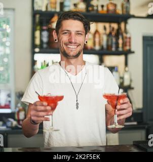 Gemischt nur für Sie. Porträt eines glücklichen jungen Barkeepers, der zwei Cocktails hält, während er hinter der Bar steht. Stockfoto