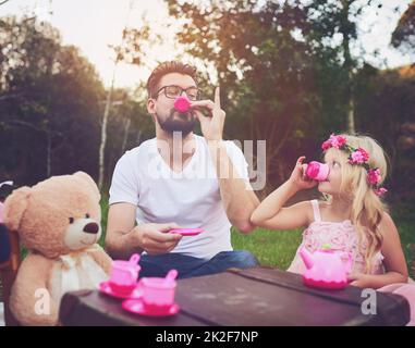 Dieser Tee ist großartig. Aufnahme einer fröhlichen Tochter und eines Vaters, die mitten in einem Garten eine Teeparty mit einem Haufen ausgestopfter Spielzeuge machen. Stockfoto