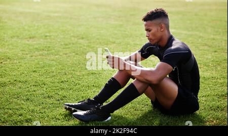 Das WiFi ist so heiß wie seine Fähigkeiten. Aufnahme eines jungen Mannes, der auf dem Feld sitzt und ein Smartphone bei einem Rugby-Spiel verwendet. Stockfoto